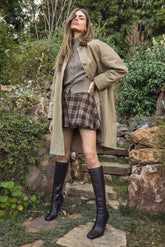 A woman is wearing a beige trench coat over a gray button-up shirt and a gray vest. She has on high-waisted, brown plaid shorts paired with knee-high black leather boots. The setting appears to be outdoors in a garden with rocks, plants, and trees in the background.